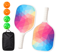 Two white-edged pickleball paddles printed with a pastel rainbow triangle pattern and wrapped white grips, shown with two orange balls, two green balls and a black carry case.